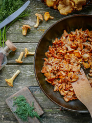 Fried chanterelle mushrooms with onion in frying pan on wooden background. Top view.A knife, a pan of mushrooms and fresh herbs