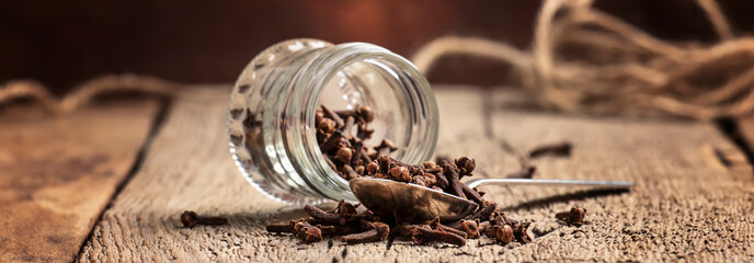 Cloves sticks in silver spoon and glass jar, old wooden table background, banner, selective focus