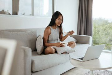 Woman having breakfast while using computer at home in morning