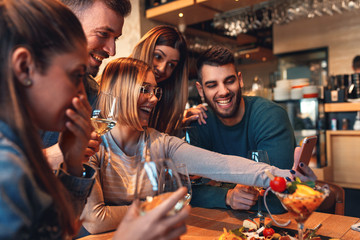 Group of young friends having fun in restaurant, they are looking at smartphone and making selfie.