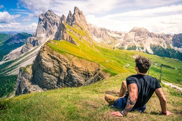 Naklejka na ściany i meble beautiful young muscular man sitting on the grass enjoying the beautiful dolomites mountains landscape. seceda, 2500m over the odle group mountains