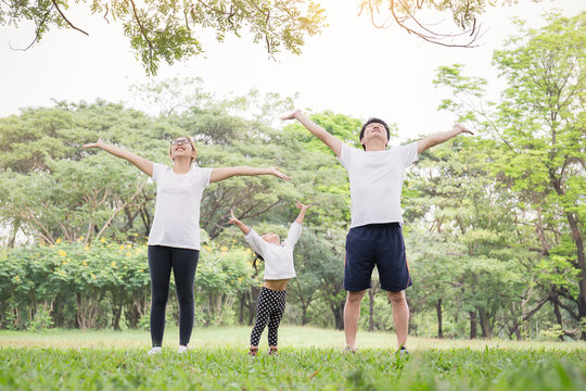 Family Exercising And Jogging Together At The Park. Asian Family Father Mother And Daughter Stretching After Sport On The Grass. Sport Health Care And Medical Freedom Relax Concept.