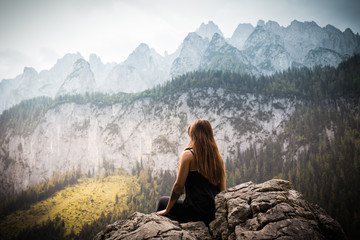 Naklejka na ściany i meble young girl with long hair sitting on the top of a rock enjoying the beautiful view of the mountains covered by forest