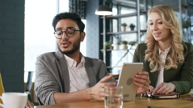 Young middle eastern businessman sitting at cafe table with blonde businesswoman, using digital tablet and talking with colleagues