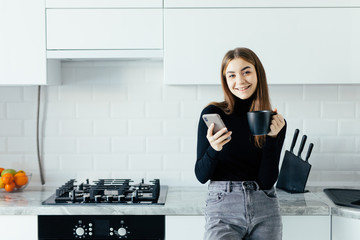 Young woman using cell phone and having a coffee in the kitchen