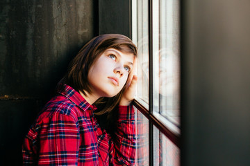 Portrait of thoughtful sad caucasian teenager boy with long straight hairstyle near the window in...