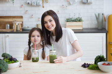 smiling mother with child daughter drinks healthy green smoothie with straw in a jar mug in kitchen. Healthy Lifestyle.