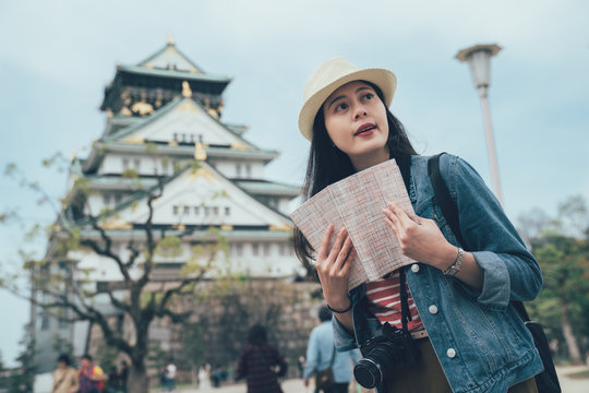Young asian female tourist traveling with paper map in japanese monument old palace on background. smiling woman traveler with camera looking aside visiting osaka castle in japan standing blue sky