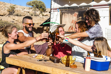 Alternative young group of friends people toasting and clinking beer bottles all together having fun in friendship in rural camping location for vacation and nature lifestyle