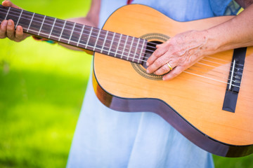 Close up of old aged senior female hands playing a guitar outdoor with green bokeh in background-...