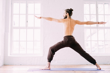 Young sporty man doing yoga exercise in white studio with wooden floor and big windows. Man and...