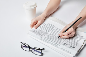 cropped view of woman holding pencil near newspaper, glasses and paper cup on white