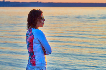 A red-haired, middle-aged woman after a wake-surfing class stands on the shore of the lake and watches the sun go down.