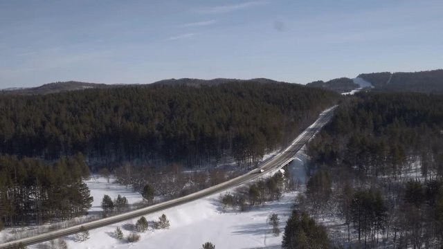 One silver minivan driving along a slightly snowy asphalt freeway through a dense forest in hilly lanscapes. Winter, sunny frozen day - Aerial drone view