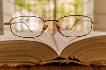 An open book on table and a specs or eyeglass on wooden table, on a warm sunny morning, side view close up. Education or Holiday Concept. Isolated background in selective focus, shallow depth of field
