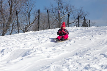 Fototapeta na wymiar Pretty smiling little girl in her ski suit sliding down a small snow covered hill with her sledge board during a bright sunny winter day