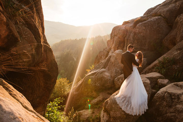 Beautiful shot of romantic wedding couple of groom and bride in gorgeous wedding dress hugging near the big rocks, cliffs in mountains on sunny day. Sunset