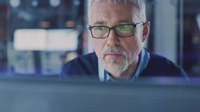 Portrait of Handsome Middle Aged Engineer Wearing Glasses Works on Personal Computer. In the Background High Tech Engineering Facility