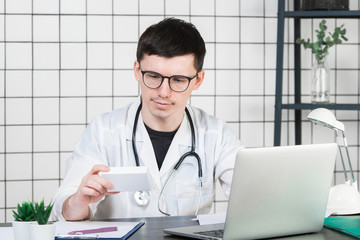 Male doctor in white coat with stethoscope over his neck sitting at table thinking on prescription, writing something down, with box of medicine in his hand
