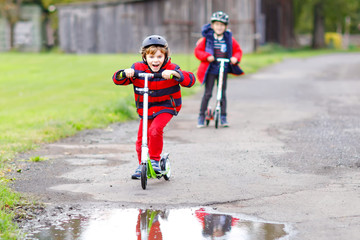 Two little kids boys riding on push scooters on the way to or from school. Schoolboys of 7 years driving through rain puddle. Funny siblings and best friends playing together. Children after school