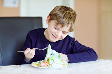 Happy kid boy eating fresh salad with tomato, cucumber and different vegetables as meal or snack. Healthy child enjoying tasty and fresh food at home or at school canteen.