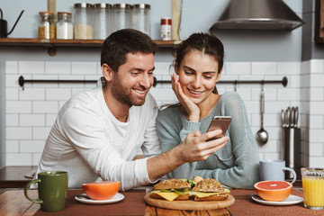 Happy young loving couple at the kitchen have a breakfast using mobile phone.