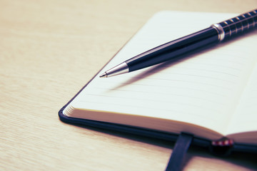 Black pen lies on the opened note cover on the spring on a light wooden table. Place to write.
