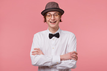 Horizontal portrait of joyful nice young guy in white shirt, hat and black bowtie wears glasses...