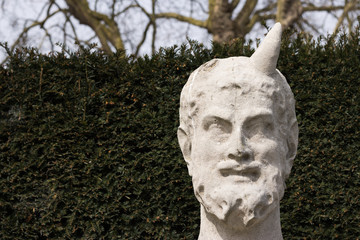 Old Bust of Horned Greek God is on Right Side of Nature Background Green Wall Outside. One Horn is Missing. Copy Space is on the Left.
