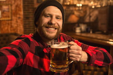 Cheerful handsome young man taking a selfie while drinking beer at the bar