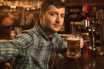 Cheerful handsome young man taking a selfie while drinking beer at the bar