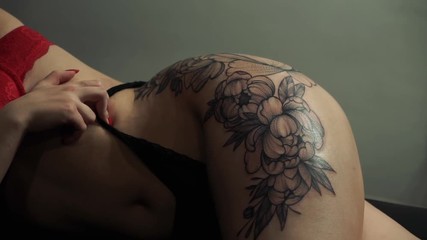 closeup young slim woman with red manicure and bra shows flower tattoo on left thigh lying in fashion studio