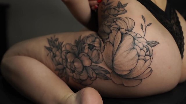 young woman in red bra and black underpants shows large peony tattoo on left thigh sitting in tattoo studio