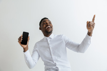 Indoor portrait of attractive young black african man isolated on grey background, holding blank smartphone, smiling at camera, showing screen, feeling happy and surprised. Human emotions, facial