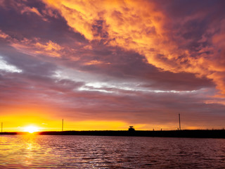 Silhouette of a tractor riding along the bank of a small river in the evening while bautiful colorful clouds are glowing in the sky and the sun is setting behind the horizon