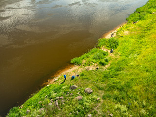 Abstract view at the bank of a river with several fishermen trying to catch some fish with their rods. Riverbank is covered with fresh green grass illiminated by summer sun