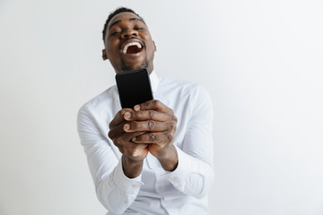 Indoor portrait of attractive young black african man isolated on grey background, holding blank smartphone, smiling at camera, showing screen, feeling happy and surprised. Human emotions, facial