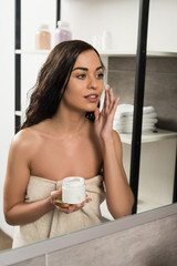 attractive woman applying face cream on cheek while looking in mirror in bathroom