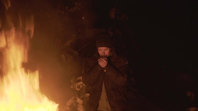 Poor man warms hands at campfire in night forest. 4K