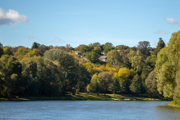 Beautiful rural landscape of blue water of river, blue sky with white clouds and several old wooden houses hiding among old huge trees in wood. Countryside of Ukraine. Horizontal color photography.