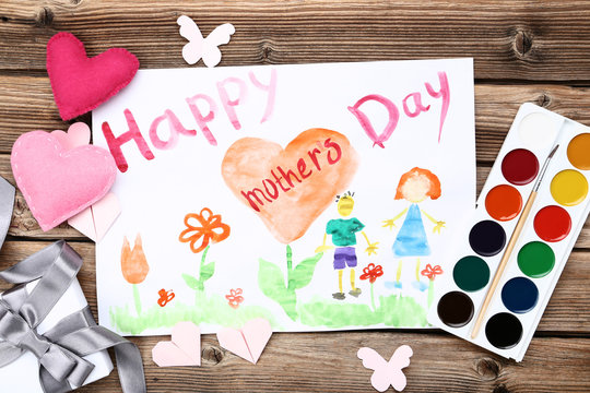 Greeting card for Happy Mothers Day with paints, gift box and hearts on wooden table