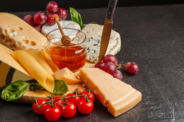 Mix cheese on dark background on wood board with grapes, honey, nuts, tomatoes and basil. Top view.