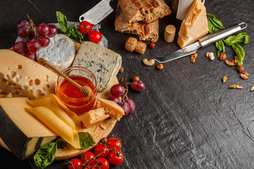 Mix cheese on dark background on wood board with grapes, honey, wine, bread, nuts, tomatoes and basil. Top view.