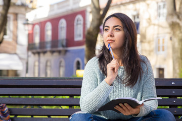 Pensive woman holding pen and copy-book on bench outdoors