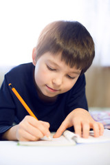 Portrait cute, cheerful little boy in a bright room. Child writing in a notebook with a pencil. Preteen schoolboy doing his homework.