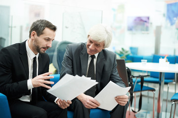 Portrait of two senior business people discussing documents sitting in office or lobby, copy space