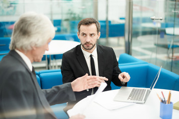 Portrait of two mature business people discussing deal sitting at table during meeting in office or lobby, copy space