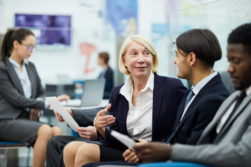 Portrait of smiling mature businesswoman talking to employees sitting on sofa in office, copy space