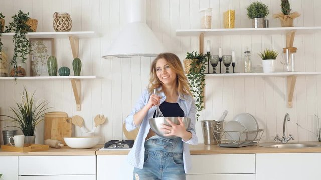 young blond woman whisks in metal bowl on table and dance happily against kitchen utensils on shelves table and stove