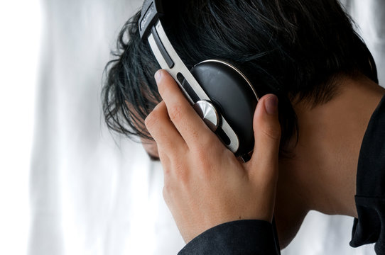 The Young Guy With An Average Hairstyle In A Dark Jacket Put On Earphones The Head And Holds Them In Hand On The White Background. He Listens To Music In Monitor Earphones And It Is Pleasant To Him.